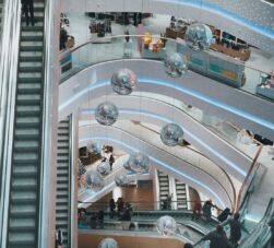 Vista de cima de shoppings em BH, onde se vê corredores com pessoas passando, escadas rolantes e grandes lustres prateados.