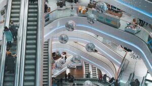 Vista de cima de shoppings em BH, onde se vê corredores com pessoas passando, escadas rolantes e grandes lustres prateados.