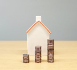 Casa em miniatura ao fundo com três pilhas de moedas em crescimento à frente, representando valorização e investimento em fundos imobiliários.