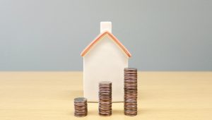 Casa em miniatura ao fundo com três pilhas de moedas em crescimento à frente, representando valorização e investimento em fundos imobiliários.